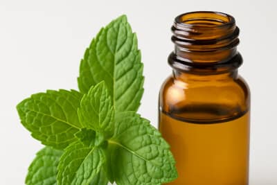 A small amber glass bottle of peppermint essential oil placed beside fresh green peppermint leaves.